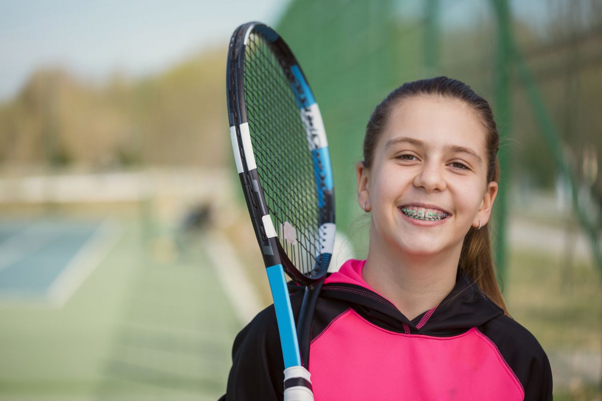 Young girl with braces smiling while holding a tennis racket on an outdoor court.